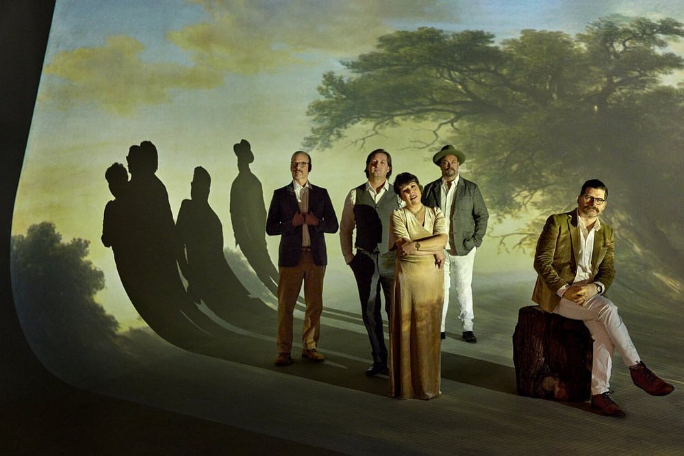 Group portrait of five people posing indoors in formal wear against a backdrop painting of a landscape with trees and shadows.