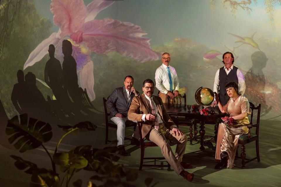 Group of six adults posed in a staged indoor scene with a floral backdrop, vintage table, and world globe.