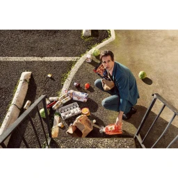Man kneeling near spilled groceries on asphalt by stairs in a parking lot by Holly Andres