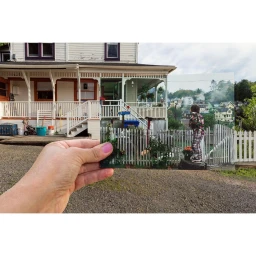 A hand holds up a small photographic print aligning with a two-story white house and picket fence in New York.