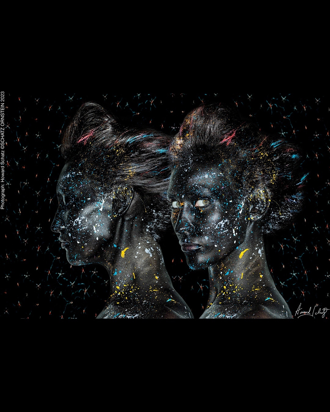 Two striking women are adorned with black body paint and vibrant paint splatters against a cosmic black background in this artistic portrait.