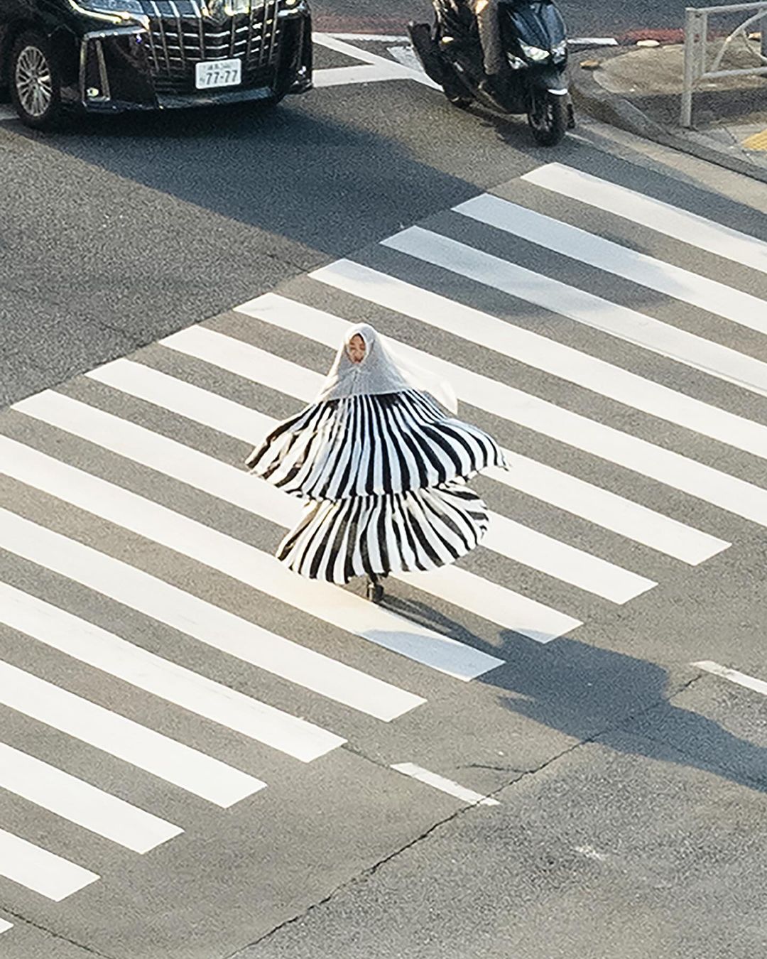 Fashionable person wearing a black and white striped dress walking on a crosswalk. A black car and scooter are in the background.