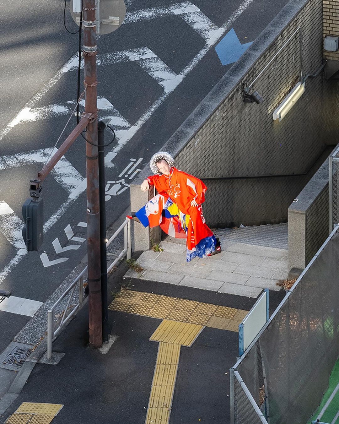 Person in orange kimono poses on staircase with colorful patterned fabric accents, urban setting, city street background, broad daylight.