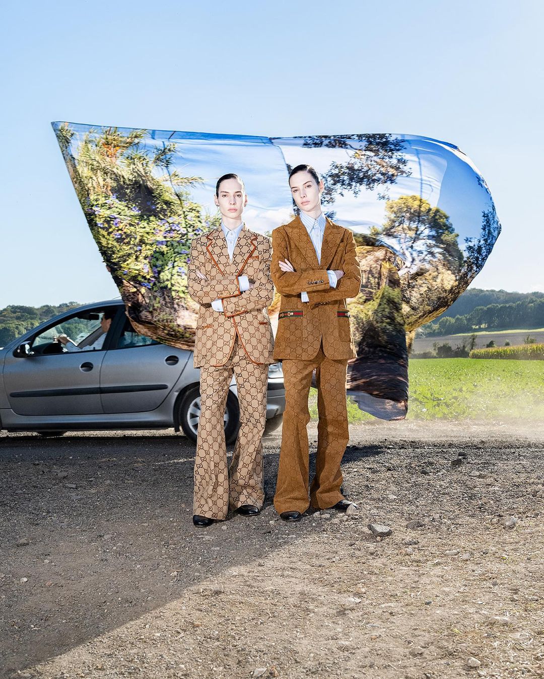 Two fashion models in matching suits pose on a dirt road next to a car and a large photographic reflector featuring a landscape.