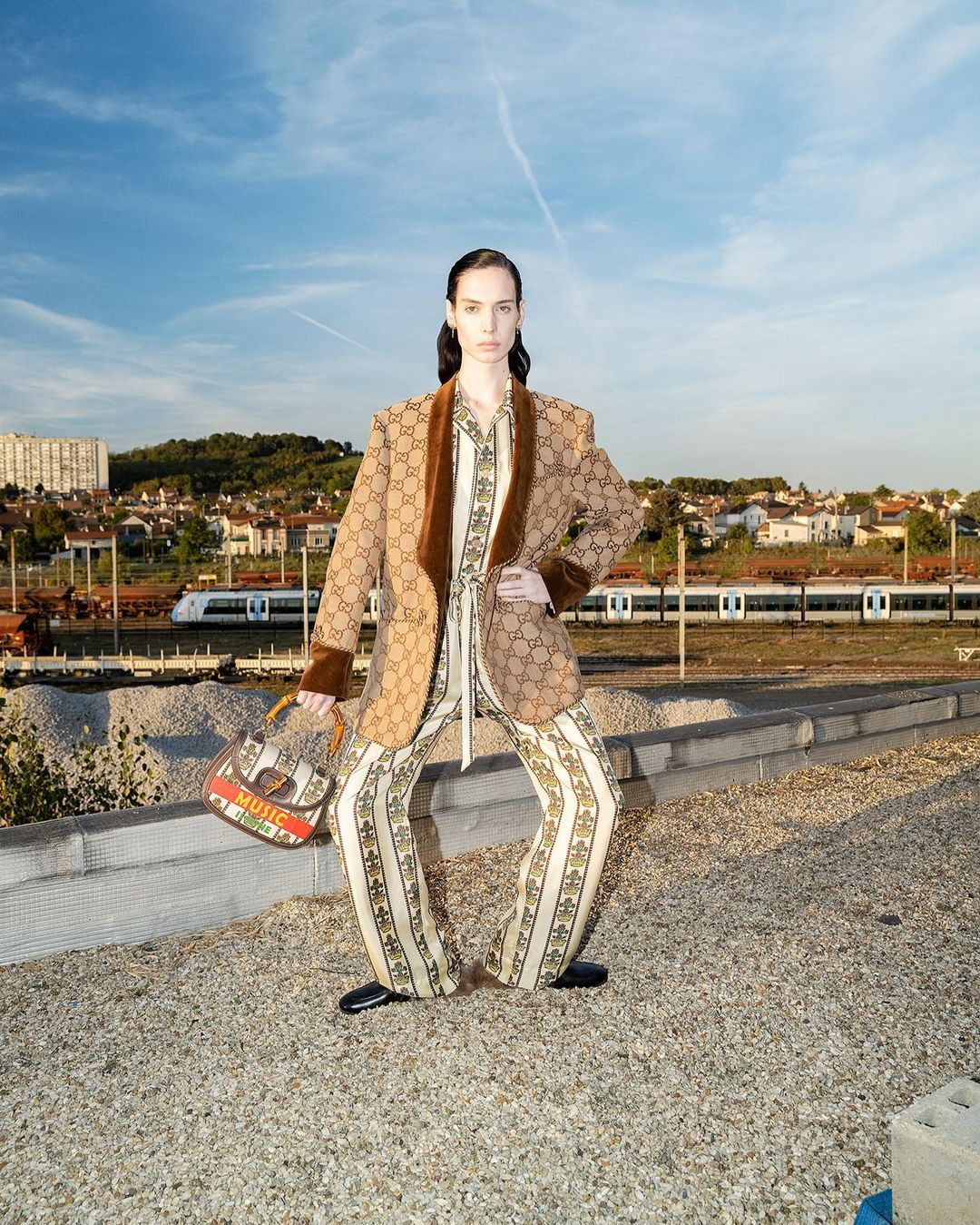 Fashion shot of a model in a patterned suit, posing outdoors on a rooftop with a handbag, with a train and buildings in the background.