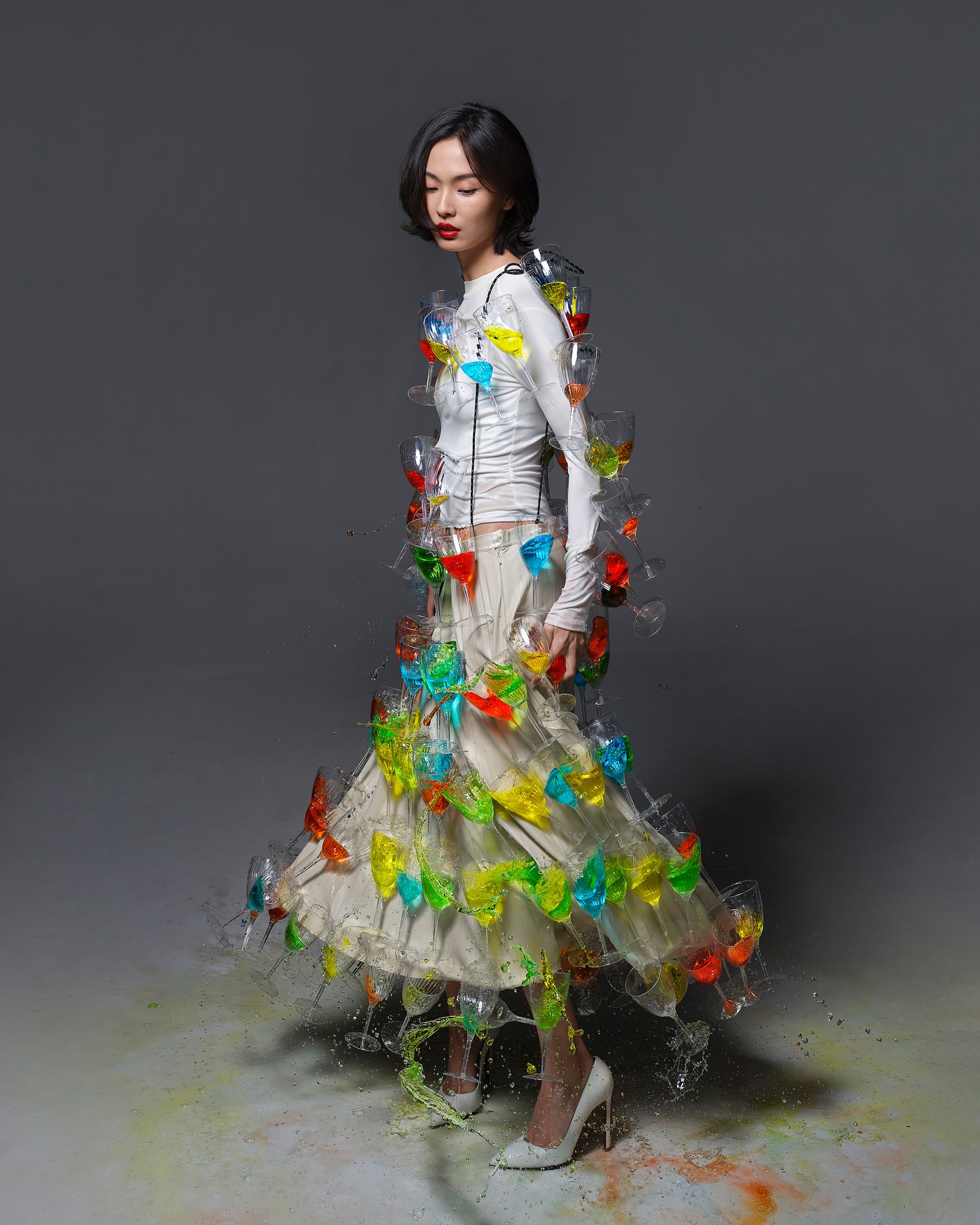 Asian woman in fashion shot wearing white top, flowing skirt, and clear wine glasses filled with color. In studio with grey backdrop.