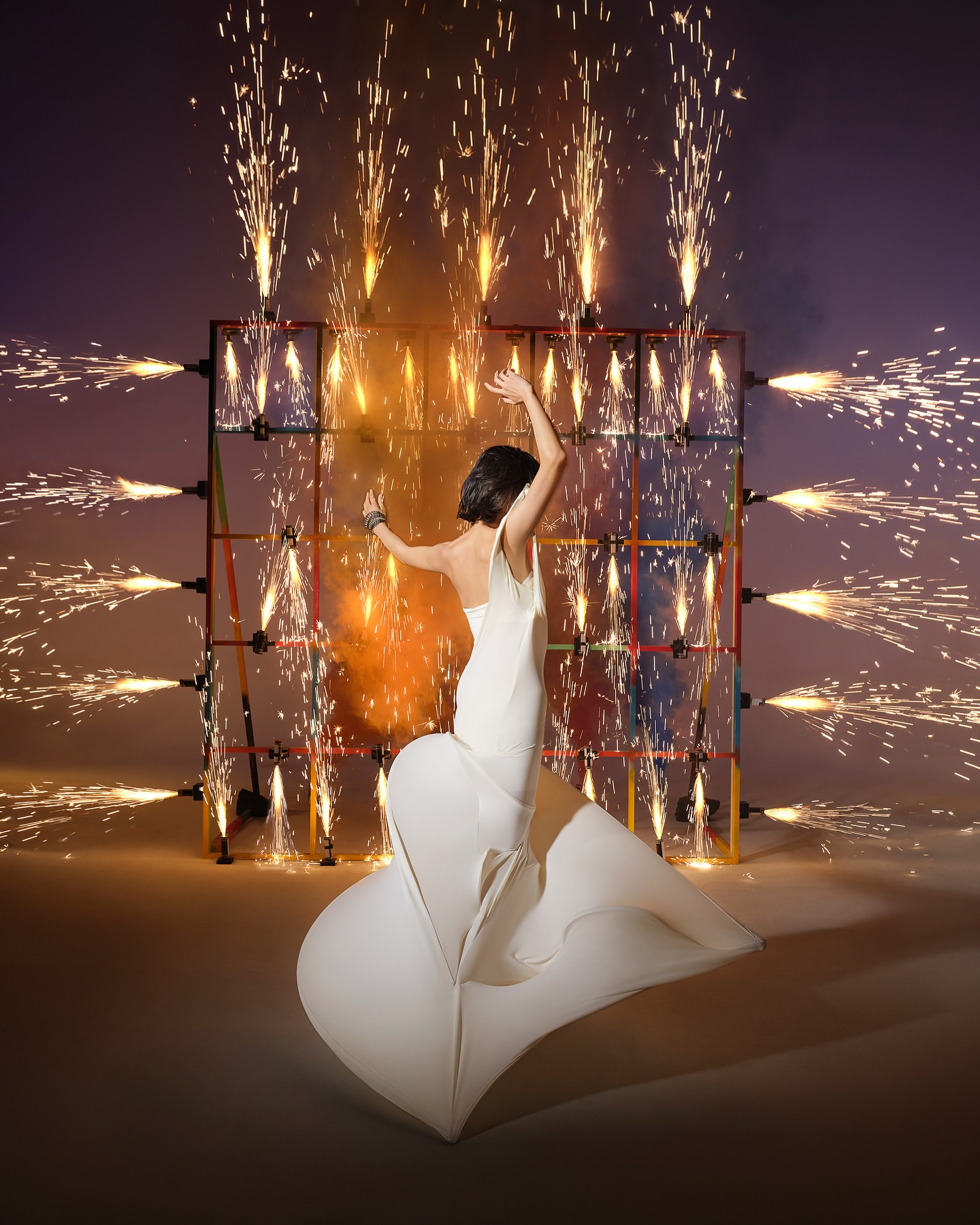 Woman in white gown poses in front of a grid of pyrotechnics, sparks flying up and out, in studio setting with purple backdrop.