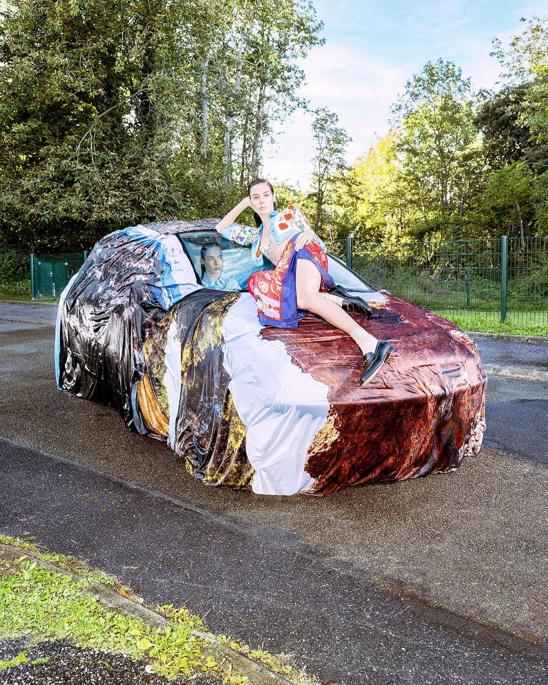Woman in colorful kimono poses on a vintage car covered with a printed tarp in an outdoor setting with trees and asphalt ground.