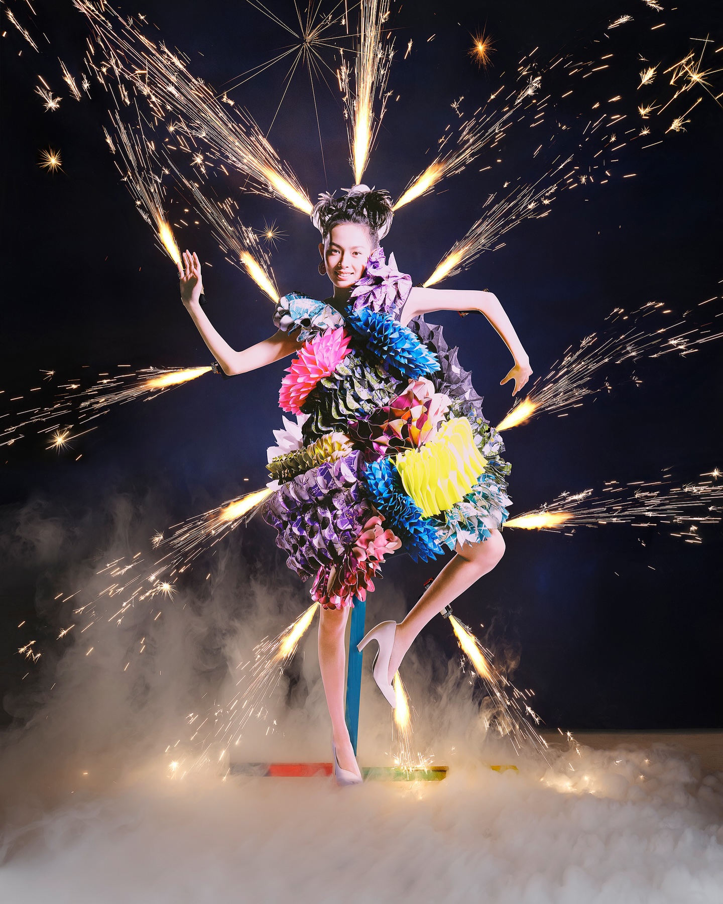 Woman poses in a creative dress made of colorful flowers and geometric elements, with a burst of fireworks and smoke in the background.