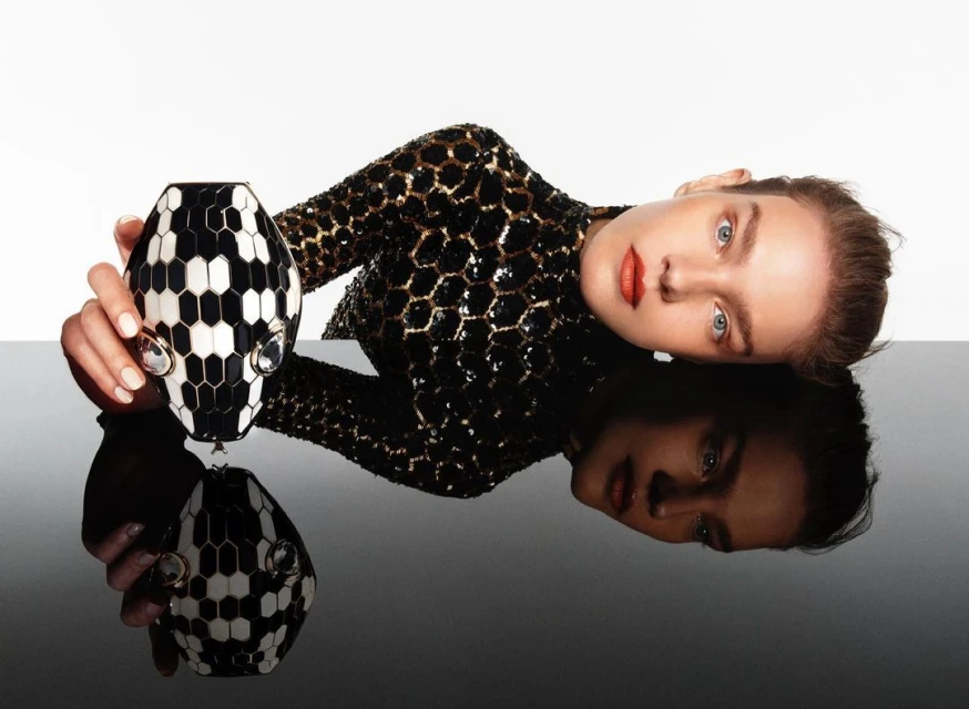 Woman in sequin dress poses with black and white clutch purse reflecting on a glossy black surface in a white studio setting.