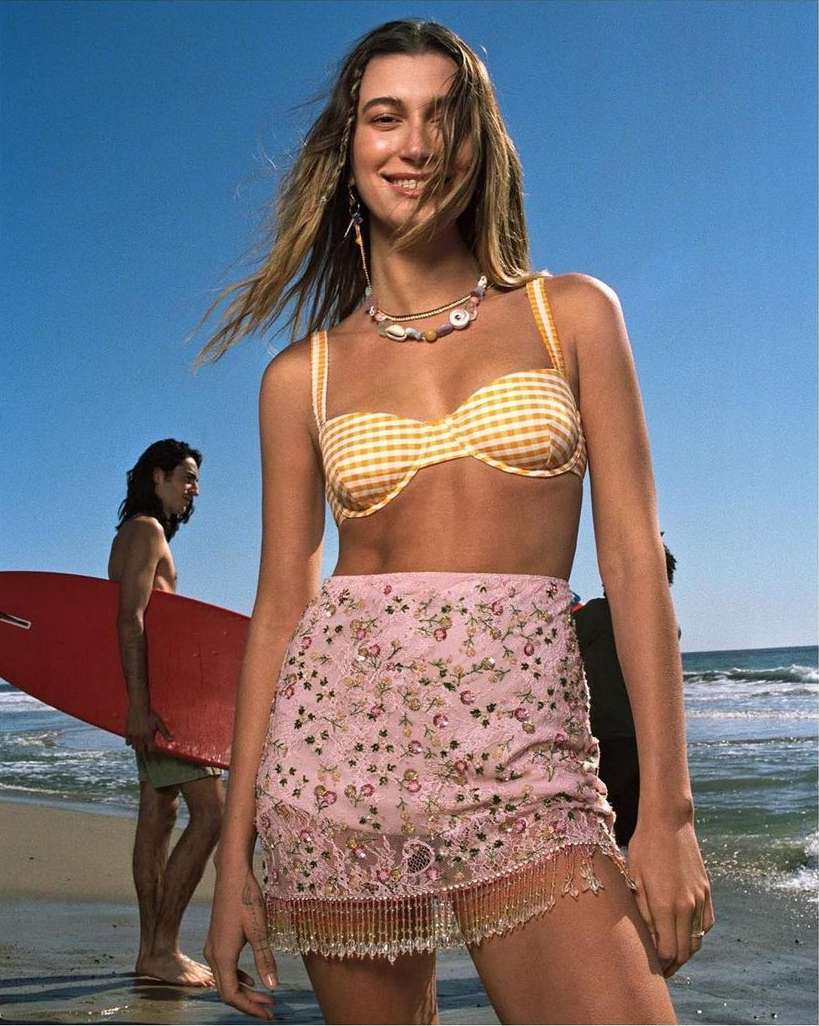 A woman smiles on the beach in a bikini top and skirt, featuring a man with a surfboard in the background against a blue sky and ocean.