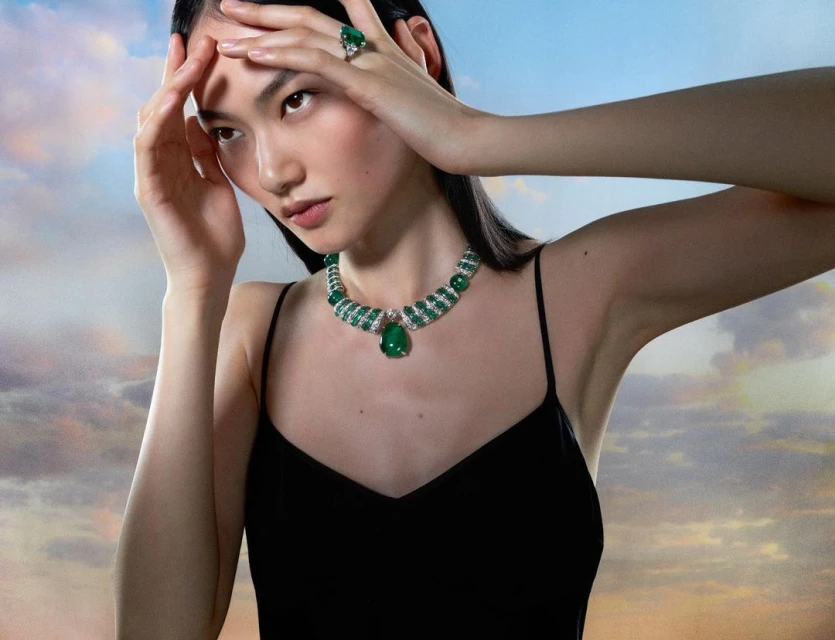 Elegant Asian woman poses in a black dress with green gemstone jewelry, framed by a soft, dreamy sky. Beauty and fashion photography.