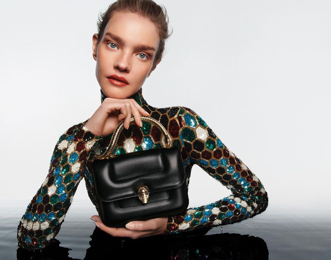 Fashion shot of woman in sequined top holding black leather handbag with gold snake detail, posing against a white and reflective surface.