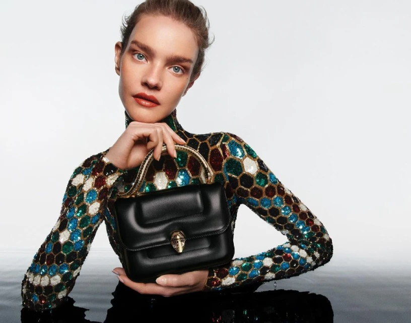 Fashion shot of woman in sequined top holding black leather handbag with gold snake detail, posing against a white and reflective surface.