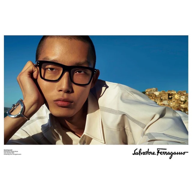 Asian male model wearing glasses and a bracelet, posing against a blue sky and cityscape backdrop for Salvatore Ferragamo.