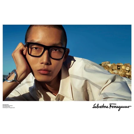 Asian male model wearing glasses and a bracelet, posing against a blue sky and cityscape backdrop for Salvatore Ferragamo.