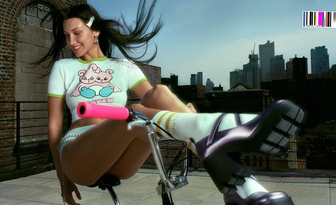 Brunette woman in a t-shirt and shorts, seated on a bicycle on a rooftop against a city skyline background.