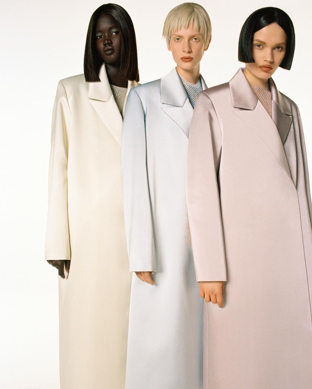 Fashion editorial featuring three women with diverse skin tones wearing long, pastel-colored coats against a bright white backdrop.