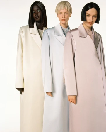 Fashion editorial featuring three women with diverse skin tones wearing long, pastel-colored coats against a bright white backdrop.