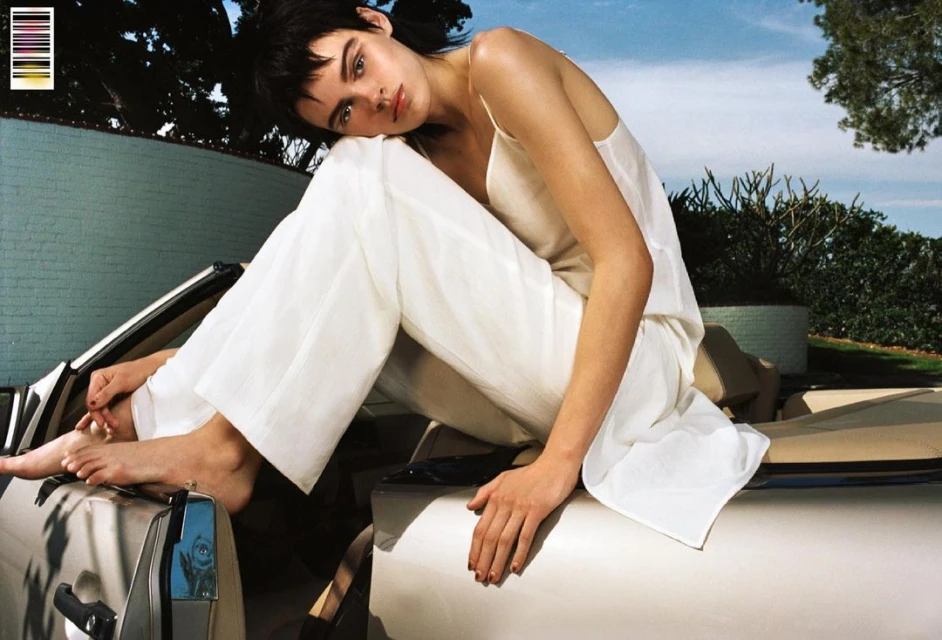 A young woman with short, dark hair in a white top and pants sits on a car. Outdoor daytime fashion shot with a blurred background.