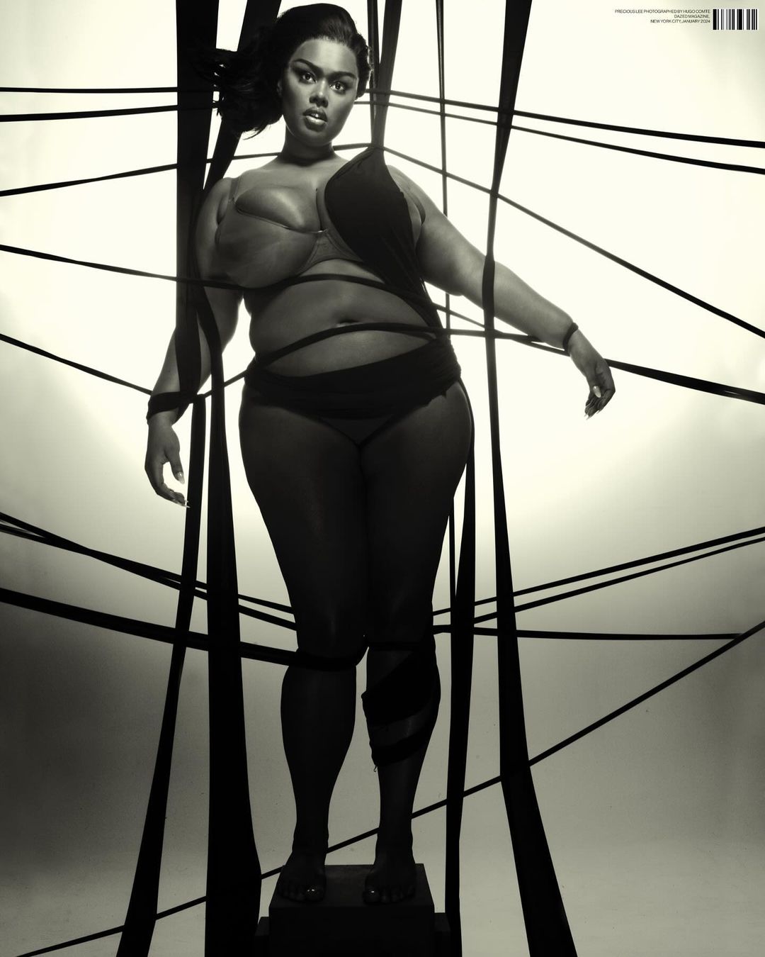 Full-figure woman in lingerie stands on a platform surrounded by black ribbons, studio shot on white background, dramatic monochrome fashion.