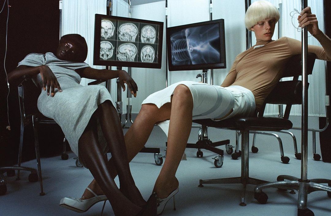 Two fashion models sit in chairs in front of medical monitors in a photo studio, showcasing minimalist fashion, with a focus on composition.