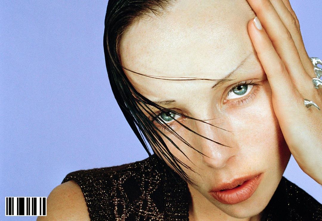 Close-up portrait of a young woman with wet hair, wearing a dark patterned top, her hand near her face, set against a lilac background.