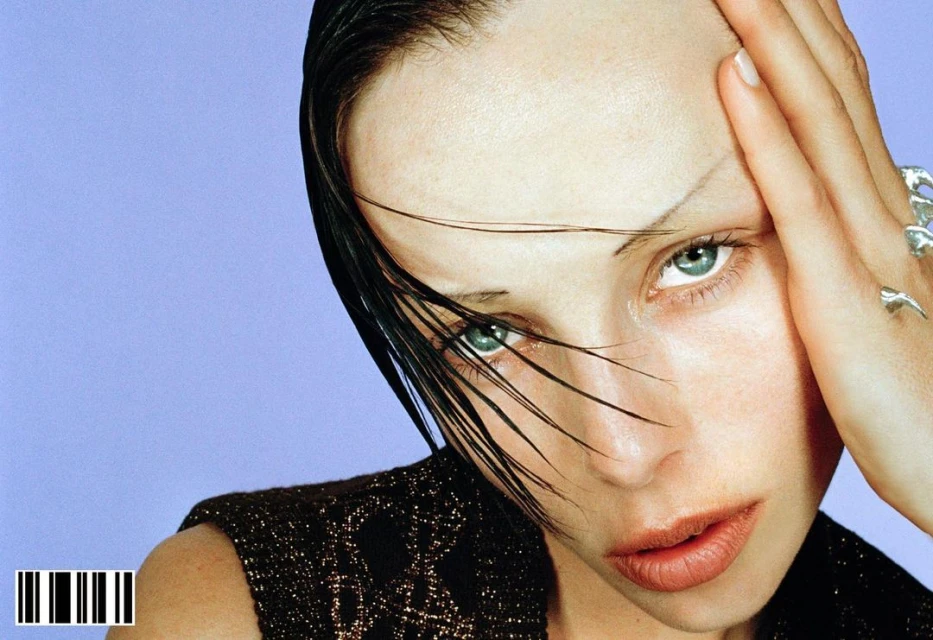 Close-up portrait of a young woman with wet hair, wearing a dark patterned top, her hand near her face, set against a lilac background.