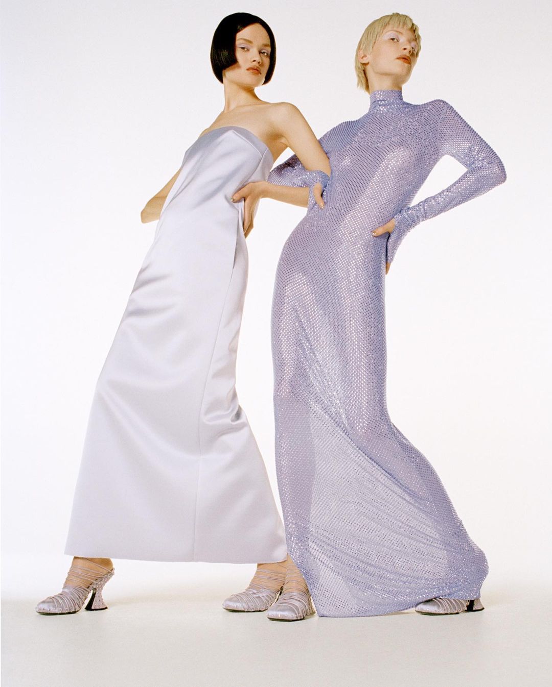 Two women in gowns pose in a studio. One wears a long satin dress; the other, a full-sleeve sequined dress. Both models are in heeled shoes.