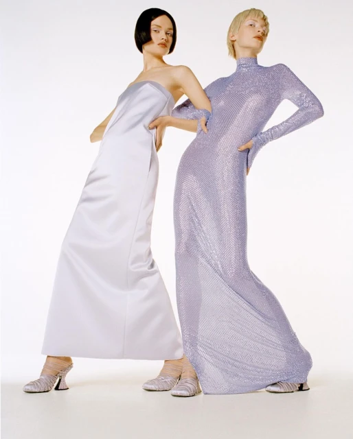 Two women in gowns pose in a studio. One wears a long satin dress; the other, a full-sleeve sequined dress. Both models are in heeled shoes.