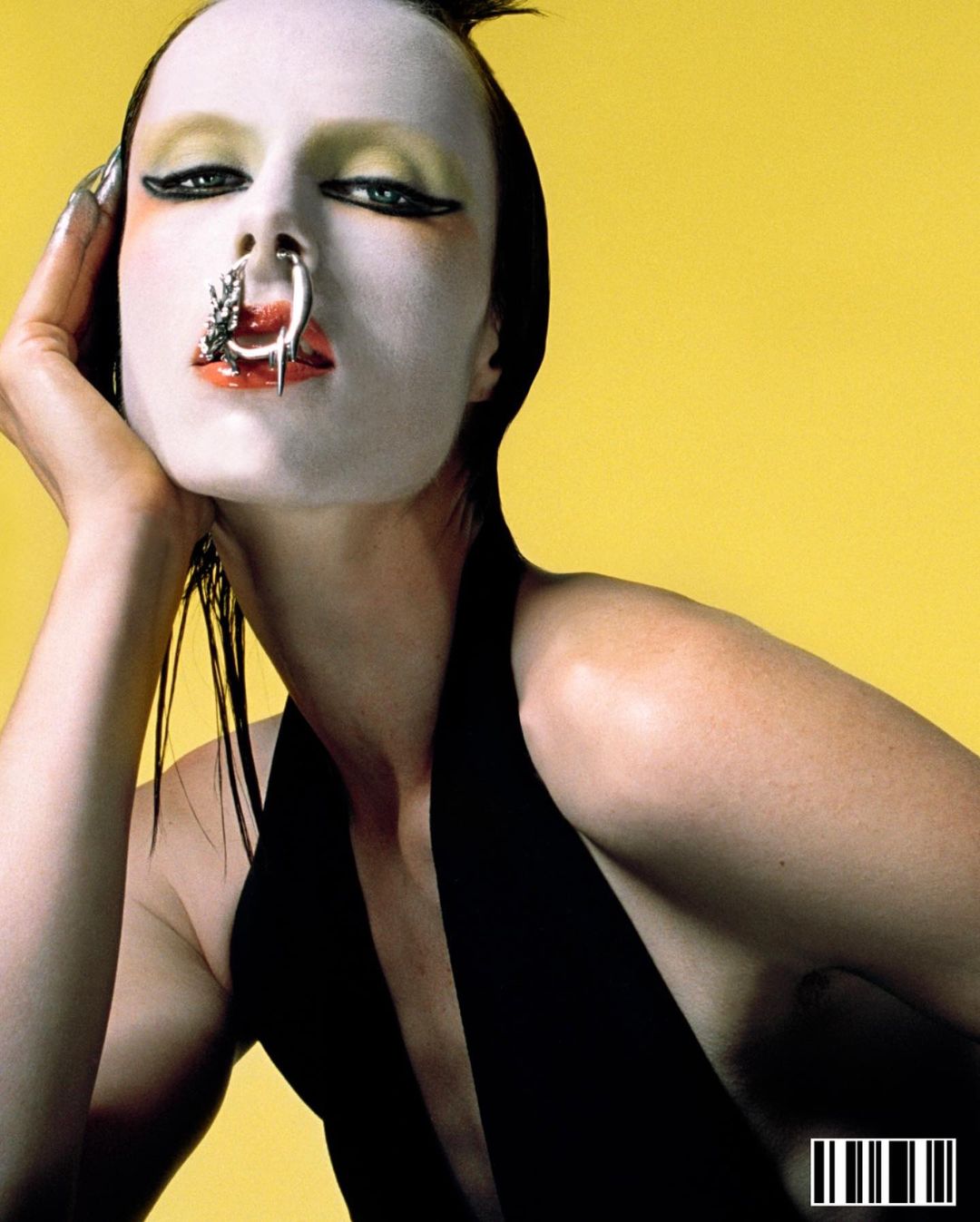 Close-up portrait of a woman with white face makeup, a nose ring, and a black top against a yellow background in a studio setting.