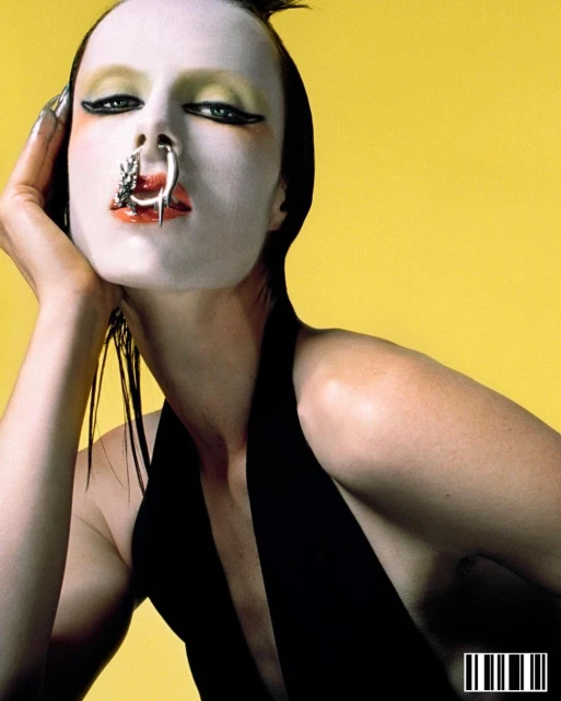 Close-up portrait of a woman with white face makeup, a nose ring, and a black top against a yellow background in a studio setting.