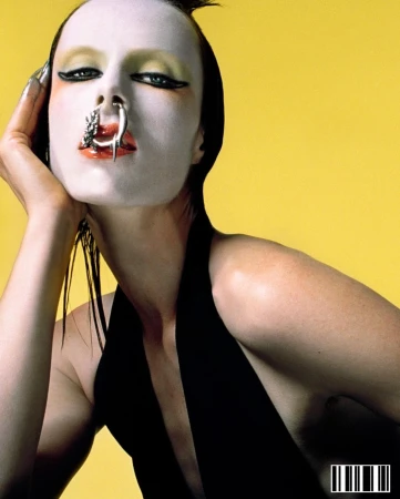 Close-up portrait of a woman with white face makeup, a nose ring, and a black top against a yellow background in a studio setting.