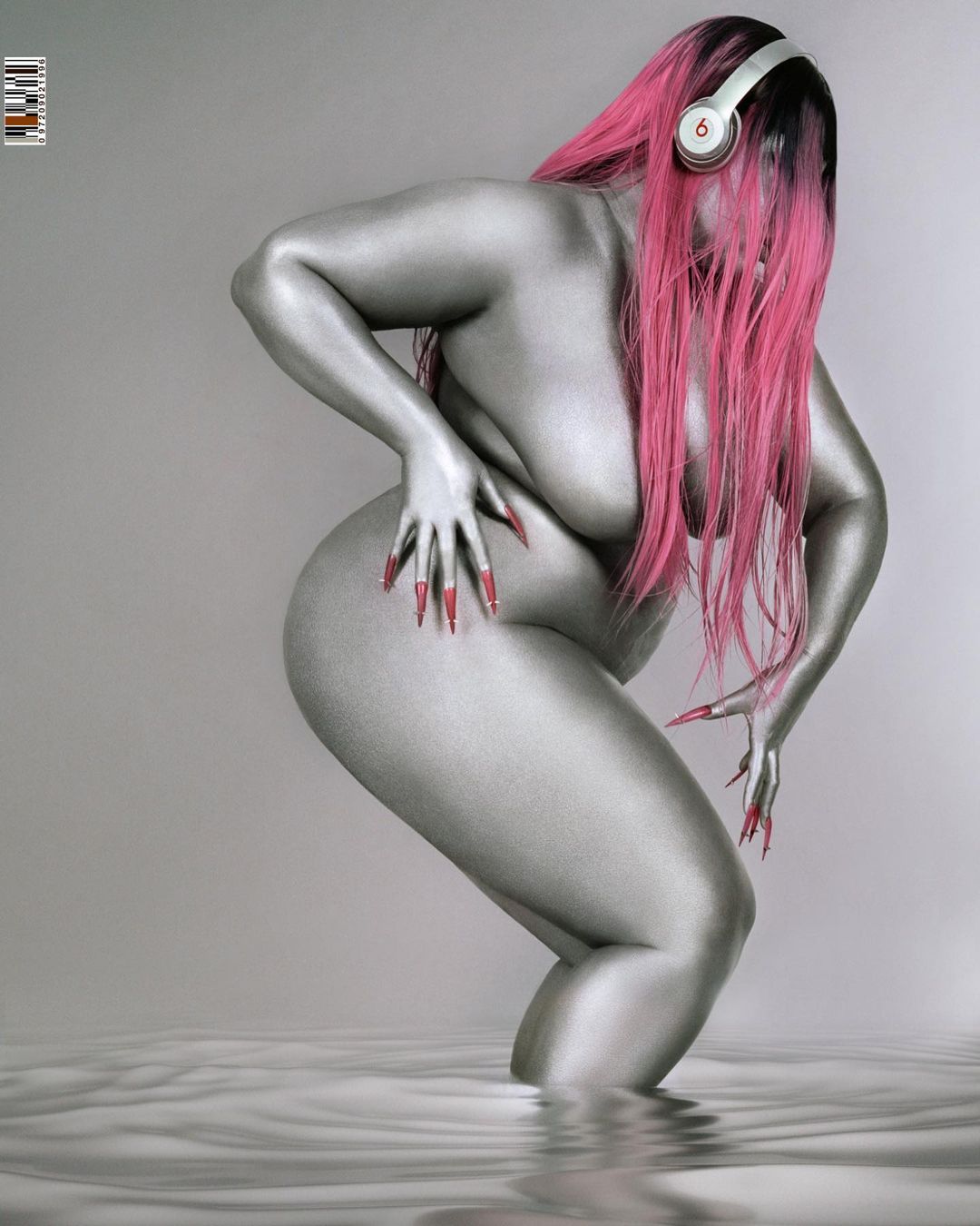 Silver painted plus-size woman with pink hair and long red nails wears headphones in a grey studio with water. Beauty shot.