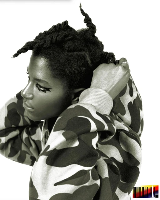 Close-up of a woman with updo hairstyle and dramatic eyeliner, wearing a camouflage hoodie against a bright white background.