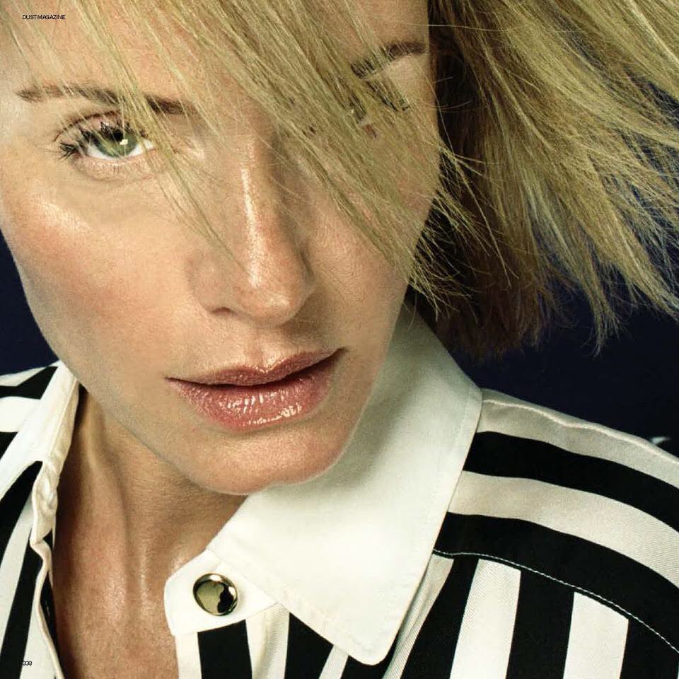 Close-up portrait of a fair-skinned adult woman with blond hair, wearing a vertically striped black and white shirt, against a dark background.