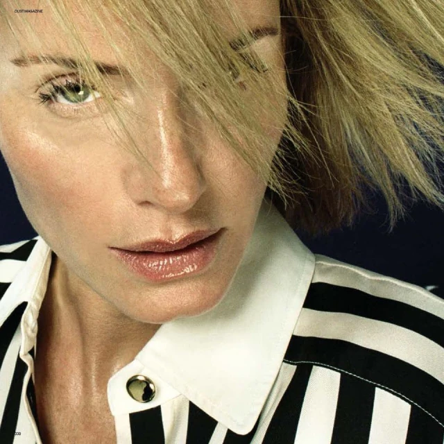 Close-up portrait of a fair-skinned adult woman with blond hair, wearing a vertically striped black and white shirt, against a dark background.