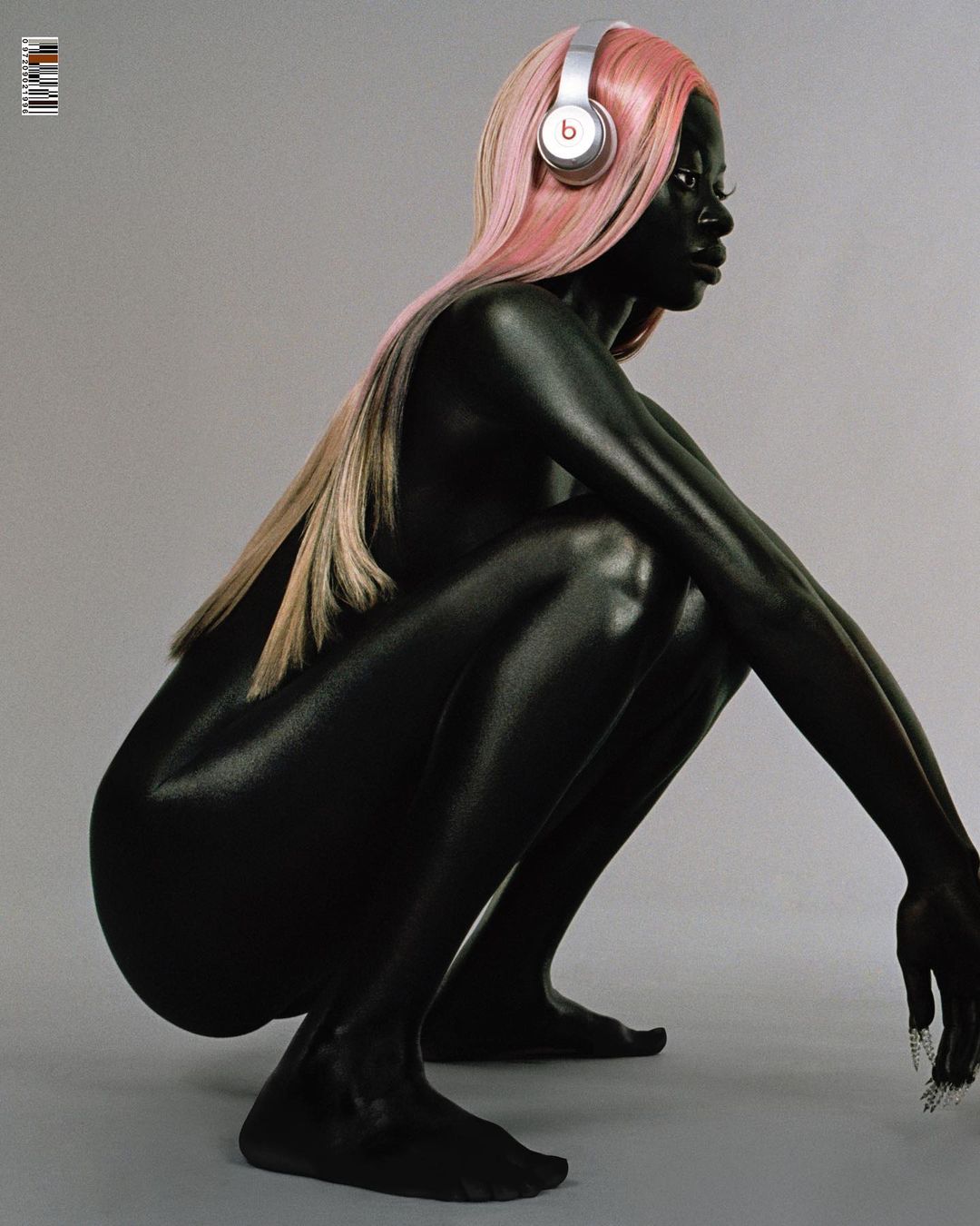 Woman with dark skin tone squats in a studio with pink hair and silver Beats headphones. The image has a plain gray background.