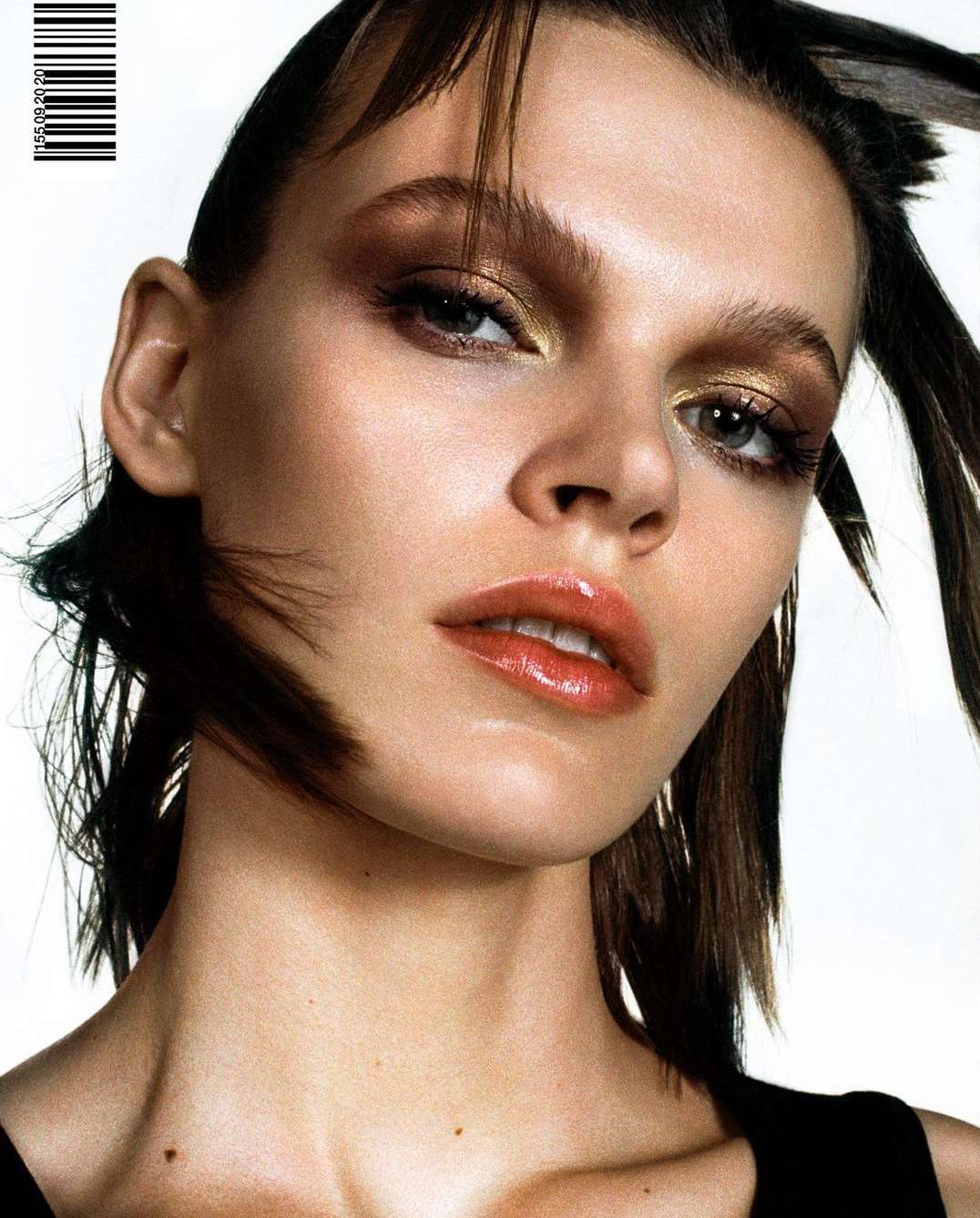 Close-up studio shot of a female model showcasing bold makeup with metallic eyeshadow, glossy lips, and a dewy complexion against a white backdrop.