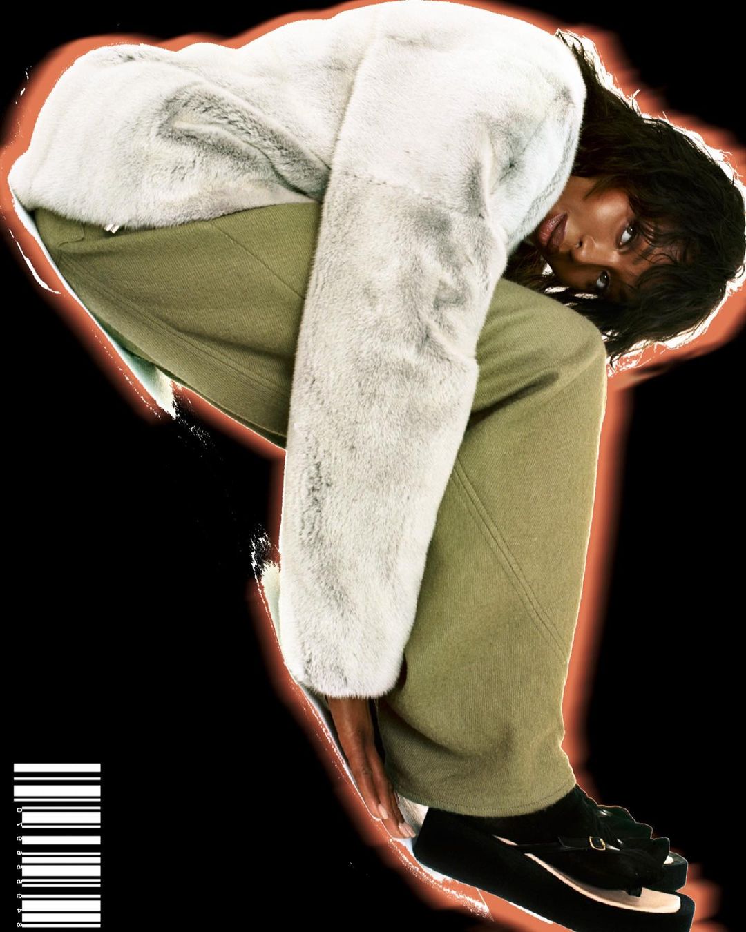 A woman models a furry grey coat and olive green pants, striking a pose against a black background with a soft orange glow.