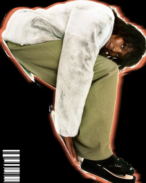 A woman models a furry grey coat and olive green pants, striking a pose against a black background with a soft orange glow.