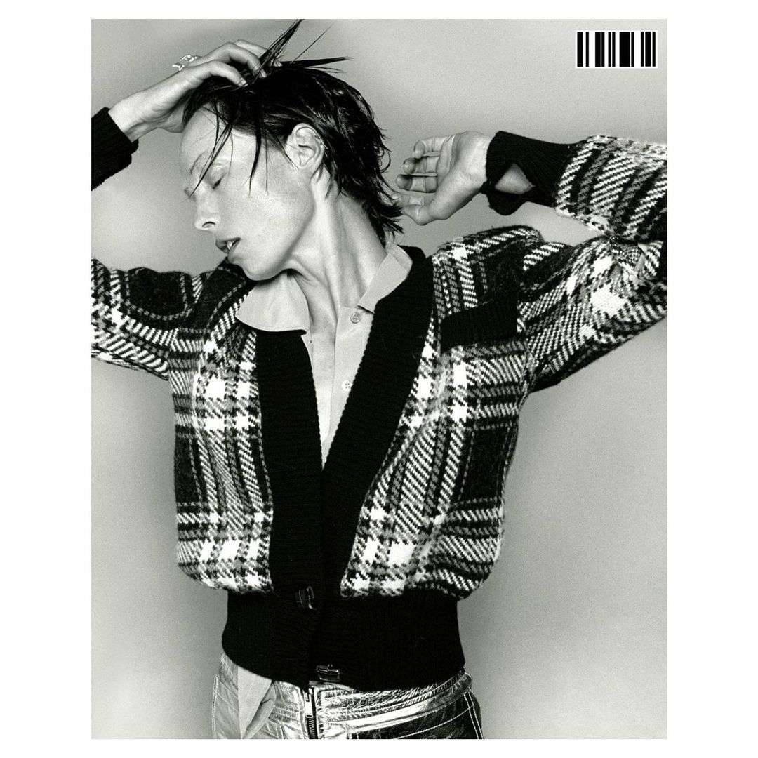 Monochrome portrait of an adult woman in plaid cardigan and metallic pants, with a wet hairstyle against a light gray backdrop.