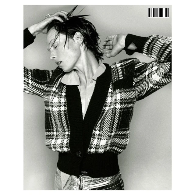 Monochrome portrait of an adult woman in plaid cardigan and metallic pants, with a wet hairstyle against a light gray backdrop.
