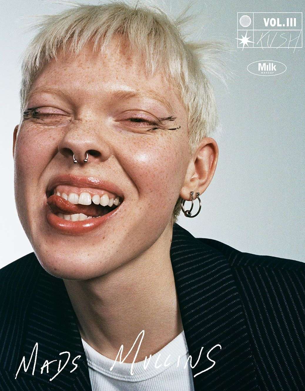 Close-up of a smiling young adult female with short blonde hair, sticking her tongue out, wearing a pinstripe blazer in a studio shot.