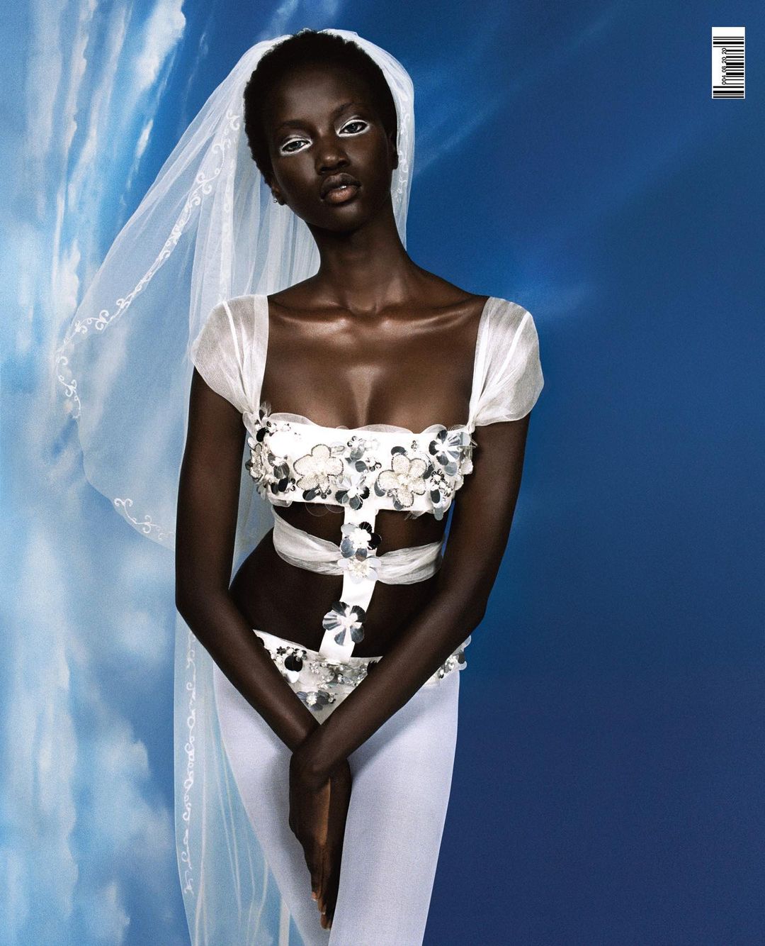 Studio portrait of an adult woman with dark skin wearing decorative white outfit and veil against a blue sky background with white clouds.
