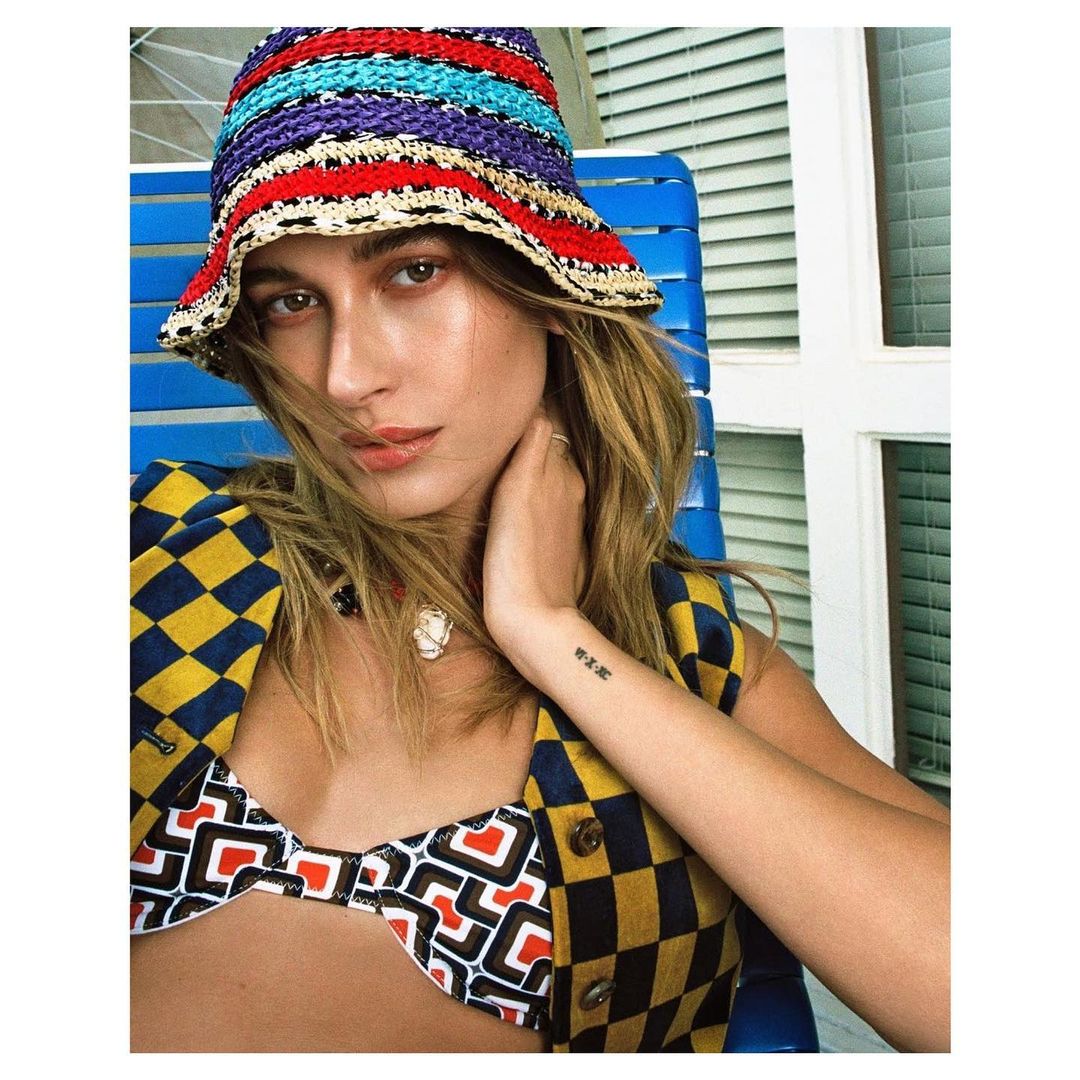 Close-up portrait of a fair-skinned woman wearing a colorful crocheted bucket hat, geometric print bikini top, and vest on a blue chair.