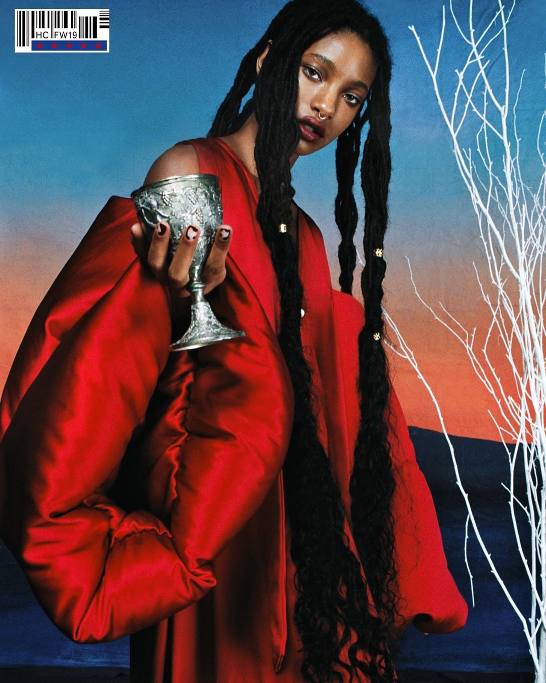 Woman in red dress holds ornate silver goblet. Model with dark hair, nose ring poses in a fashion editorial shot against dusk sky.