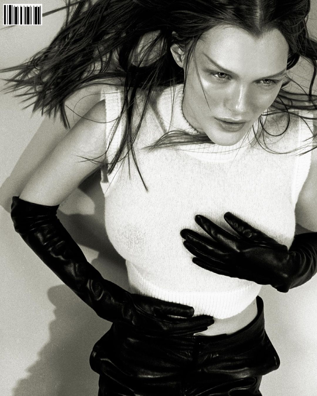 Black-and-white portrait of a fair-skinned woman with dark hair, wearing a white top and black leather gloves and pants.