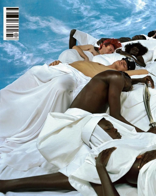 Diverse group of people in white cloth lie on a bed against a blue, cloudy sky backdrop in an indoor studio setting.