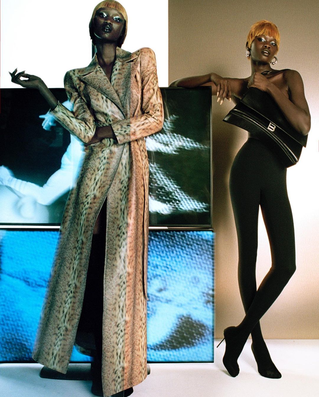 Two adult women pose in a studio setting, wearing fashionable clothing. One is wearing a printed coat and the other leggings with a bag.